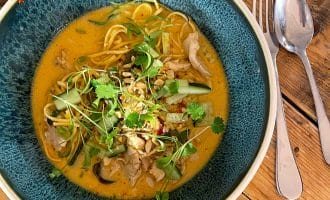 A bowl of noodle soup with sliced vegetables, herbs, and chicken in a creamy, orange broth, served on a rustic wooden table with a spoon and fork beside the bowl.