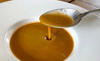 A spoon pours thick brown gravy into a shallow white bowl, with some gravy already pooled at the bottom. The scene is set on a wooden table with a blue and white cup in the background.