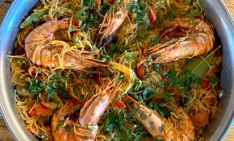 A metal pan filled with cooked noodles, large prawns, chopped tomatoes, and garnished with fresh herbs, sits on a wooden table.