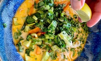 A hand squeezes a lime wedge over a bowl of colourful curry soup with noodles, leafy greens, carrots, and chopped fresh herbs in a blue dish.