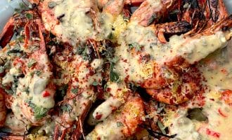 Grilled prawns with heads on, served on a white platter, topped with a creamy herb and lemon sauce, sprinkled with fresh herbs and red pepper flakes.