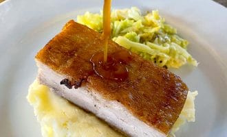 A thick slice of crispy roast pork belly sits on mashed potatoes, with gravy being poured over it. A side of cooked cabbage is also on the white plate.