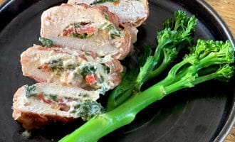 Sliced stuffed chicken breast filled with spinach, cheese, and red peppers, served on a dark plate with steamed tenderstem broccoli on the side, placed on a wooden table.