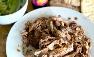 Shredded cooked meat on a white plate, with pitta bread, a bottle of sauce, and a bowl of lettuce and cucumber on a wooden table.