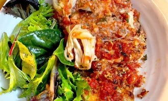 A white plate with a serving of aubergine Parmesan topped with tomato sauce and melted cheese, next to a side of mixed leafy green salad.