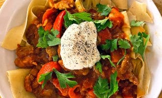 A white plate of wide pasta topped with a tomato-based meat stew, red peppers, fresh parsley, and a dollop of soured cream sprinkled with black pepper, served on a wooden table.