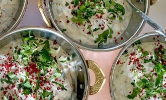 Four metal bowls with ornate handles are filled with creamy raita, garnished with chopped fresh herbs and red spices. Spoons rest in each bowl, and the bowls are arranged closely together on a light surface.