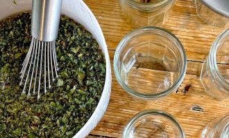 A metal whisk in a bowl of chopped green herbs sits on a wooden table next to several empty glass jars and a metal funnel, ready for filling.