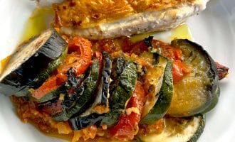 A plate with a sliced, golden-brown roasted chicken breast and a serving of colourful ratatouille made with courgette, aubergine, and tomatoes, all arranged neatly on a white dish.
