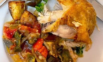 A plate with a roasted chicken leg, roast potatoes, and a colourful mix of cooked vegetables, including tomatoes, peppers, and aubergine, with a fork and knife on the side.