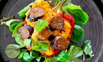 A halved red pepper filled with mashed potato and sliced sausage, garnished with fresh herbs and parsley, served on a dark plate with leafy greens underneath.