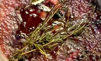 A close-up of a simmering pot with red wine, fresh thyme sprigs, a bay leaf, and chunks of vegetables, likely preparing a sauce or reduction. Bubbles are visible on the surface as it cooks.