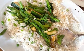 A white oval plate with steamed white rice topped with green vegetables, including sprouting broccoli, mangetout, and courgette, garnished with sliced spring onions and red chilli flakes. A serving spoon rests on the plate.