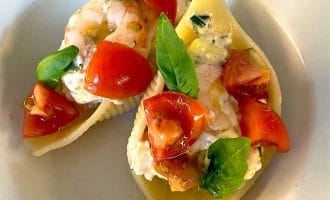 Two pasta shells filled with creamy cheese, prawns, chopped tomatoes, and fresh basil leaves, served on a white plate.