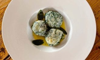 Three spinach and ricotta dumplings served on a white plate with sage leaves and melted butter, placed on a rustic wooden table.