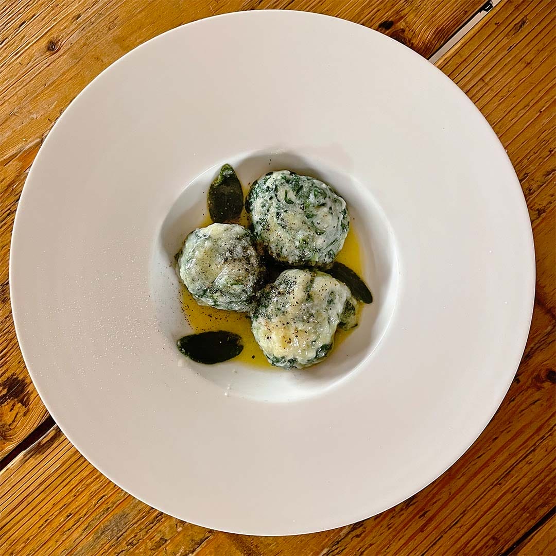 Three spinach and ricotta dumplings served on a white plate with sage leaves and melted butter, placed on a rustic wooden table.
