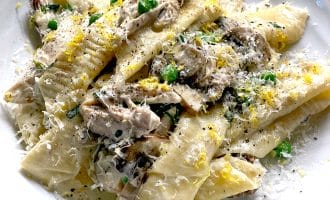 A plate of wide, homemade pasta with creamy mushroom sauce, topped with grated cheese, chopped herbs, and lemon zest, served on a white dish.