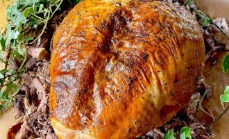 A golden-brown, roasted turkey breast is surrounded by shredded turkey meat and garnished with fresh green herbs on a wooden surface.