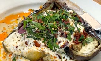 A roasted aubergine half topped with creamy yoghurt sauce, chopped fresh herbs, red pepper flakes, and a drizzle of oil, served on a white plate with a knife and fork on a wooden table.