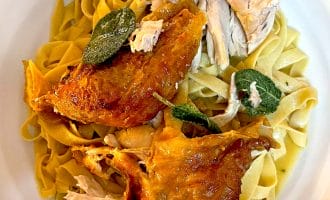 A plate of fettuccine pasta topped with roasted chicken pieces and crispy sage leaves, served on a white dish.