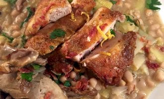 A close-up of a plate with crispy sliced chicken on top of a hearty white bean stew, garnished with fresh parsley and lemon zest. The stew contains beans, leeks, and shredded meat.