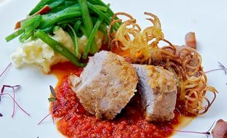 Two thick slices of cooked meat sit on a bed of red tomato sauce, served with mashed potato, green beans, crispy shoestring chips, and garnished with microgreens on a white plate.
