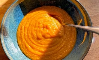 A blue ceramic bowl filled with thick, orange soup sits on a wooden table, with a spoon inside. Sunlight and shadows fall across the bowl, and a glass of water is visible in the background.