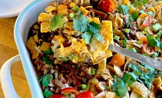 A casserole dish filled with a layered Frito pie topped with corn chips, melted cheese, minced beef, tomatoes, spring onions, and coriander. A serving is being lifted out with a spatula. A stack of white plates is nearby.