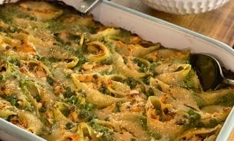 A large white baking dish filled with stuffed pasta shells topped with melted cheese and green herbs, with a serving spoon inside. In the background, there is a bowl of salad and a bottle of olive oil.