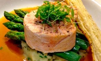 A gourmet dish featuring a round piece of salmon atop mashed potato with green asparagus spears, garnished with microgreens, served with a brown sauce and a puff pastry stick on the side.
