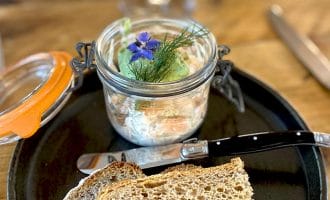 A black plate with two slices of rustic bread, a knife, and an open glass jar containing a creamy spread garnished with fresh herbs and edible flowers. The background shows a wooden table and a blurred wine glass.
