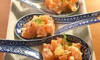 Four decorative ceramic spoons with diced raw salmon, onions, and herbs, garnished with dill and pepper, arranged neatly on a rectangular plate.