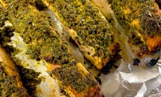 Herb-crusted salmon fillets baked on a sheet of aluminium foil, with a golden green topping and slightly crispy edges. The fish appears moist and well-seasoned.