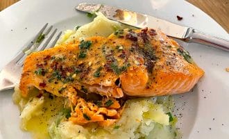 A plate of mashed potatoes topped with a seasoned, baked salmon fillet, garnished with herbs and spices. A fork and knife rest on the plate, and a glass is visible in the background on a wooden table.