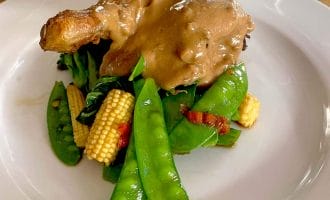 A roasted chicken leg topped with creamy brown sauce, served on a bed of sautéed mangetout, baby sweetcorn, and greens on a white plate.