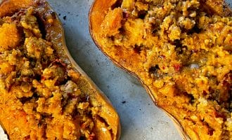 Two halves of butternut squash are filled with a crumbly, golden brown stuffing and baked on a metal tray. The stuffing appears to include grains and vegetables, giving a textured appearance.