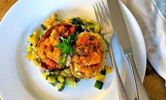 A white plate with stuffed peppers filled with a vegetable and potato mixture, garnished with parsley, and surrounded by diced sautéed courgette. A fork and knife rest at the side of the plate, on a wooden table.