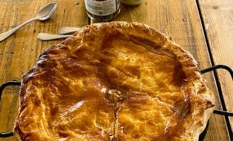 A golden-brown, freshly baked pie in a tin sits on a rustic wooden table, with two spoons and jars of condiments in the background. The pie has a flaky, glossy pastry lid and a single cut starting from the centre.