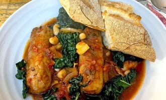 A white plate with sausage stew, featuring sausages, leafy greens, beans, and tomato sauce, garnished with two pieces of crusty bread on top. A fork and spoon rest on a serviette nearby.