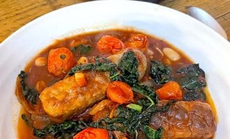 A white bowl filled with sausage stew, featuring sausages, cherry tomatoes, greens, and beans in a tomato-based sauce. A crusty piece of bread sits on a small black plate in the background.