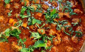A close-up of a pot filled with a chunky tomato-based stew, featuring pieces of meat, vegetables, and topped with fresh chopped herbs.