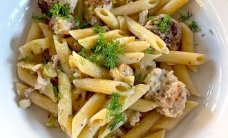 A white bowl filled with penne pasta, meatballs, creamy sauce, and garnished with fresh herbs, sitting on a wooden table.