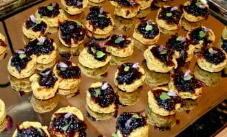A tray filled with small, round appetiser bites topped with a dark jam or chutney and garnished with tiny herbs and edible flowers, arranged neatly for serving.