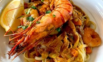 A bowl of pasta topped with a large grilled king prawn, creamy tomato sauce, chopped herbs, and a lemon wedge on the side. Prawn pieces and diced vegetables are mixed in with the pasta.
