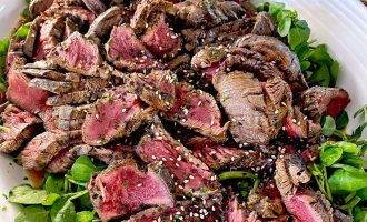 Slices of medium-rare steak topped with sesame seeds, served on a bed of fresh salad leaves on a white platter.