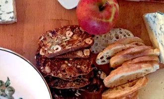 A wooden table with slices of baguette, nutty crispbread, an apple, various cheeses, and a plate with cutlery in the corner.