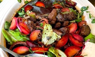 A white bowl filled with a colourful salad of leafy greens, sliced plums, seared beef pieces, and a dark balsamic dressing, with a large serving spoon resting inside the bowl.