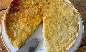 A partially sliced onion tart sits on a white plate atop a wooden table, with a metal serving spatula resting nearby. The tart has a golden-brown, slightly uneven surface and a flaky pastry.
