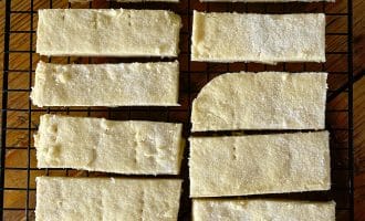 Rectangular pieces of golden, sugar-dusted shortbread are cooling on a black wire rack set over a wooden surface. The shortbread bars are arranged neatly in rows.