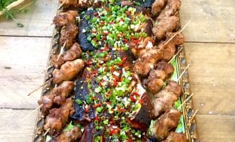 A large tray of grilled meat skewers arranged along the sides, with assorted grilled meats in the centre topped with chopped spring onions and red chillies, displayed on a wooden table.